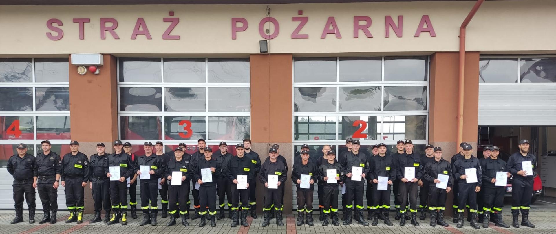 Na zdjęciu druhowie OSP stoją w dwuszeregu po zakończeniu Kursu Dowódców. W rękach trzymają zaświadczenia ukończenia kursu. Ubrani są w ubrania koszarowe. Obok nich stoją członkowie komisji egzaminacyjnej. W tle bramy garażowe budynku komendy PSP w Strzyżowie