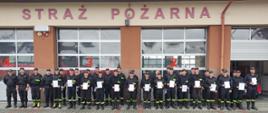 Na zdjęciu druhowie OSP stoją w dwuszeregu po zakończeniu Kursu Dowódców. W rękach trzymają zaświadczenia ukończenia kursu. Ubrani są w ubrania koszarowe. Obok nich stoją członkowie komisji egzaminacyjnej. W tle bramy garażowe budynku komendy PSP w Strzyżowie