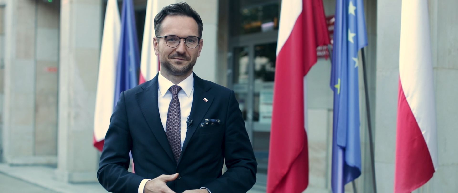 Na zdjęciu wiceminister Waldemar Buda stojący przed Ministerstwem Funduszy i Polityki Regionalnej. W tle flagi Polski i Unii Europejskiej.