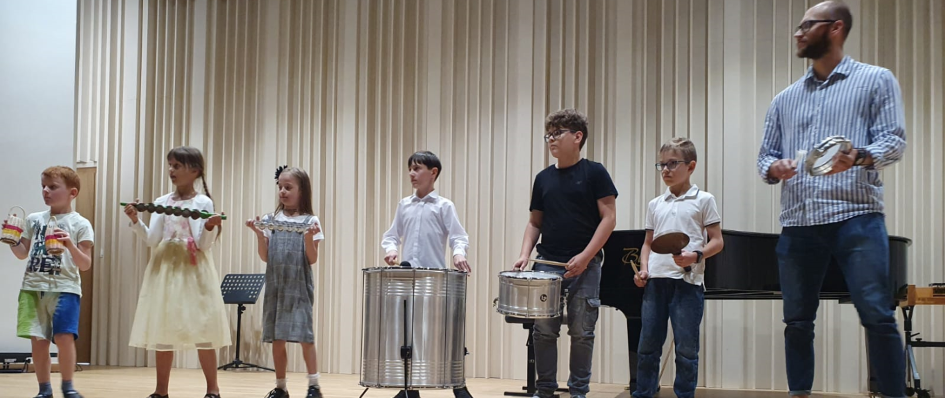 Na scenie stoi sześcioro uczniów, każdy trzyma instrument perkusyjny. Mamy czterech chłopców i dwie dziewczynki. Obok nich po prawej stronie stoi mężczyzna- nauczyciel, który także trzyma instrument perkusyjny. Mężczyzna ubrany jest w biało- niebieską koszulę i granatowe spodnie, nosi okulary i lekkich zarost. W tle za nimi stoi czarny fortepian.