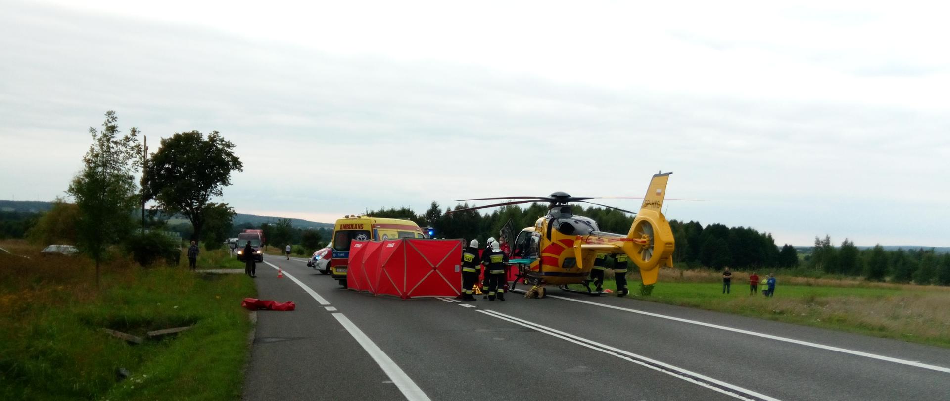 Zdjęcie przedstawia śmigłowiec lotniczego pogotowia ratunkowego który wylądował na jezdni. Obok znajduje się parawan który zasłania ofiarę wypadku.
