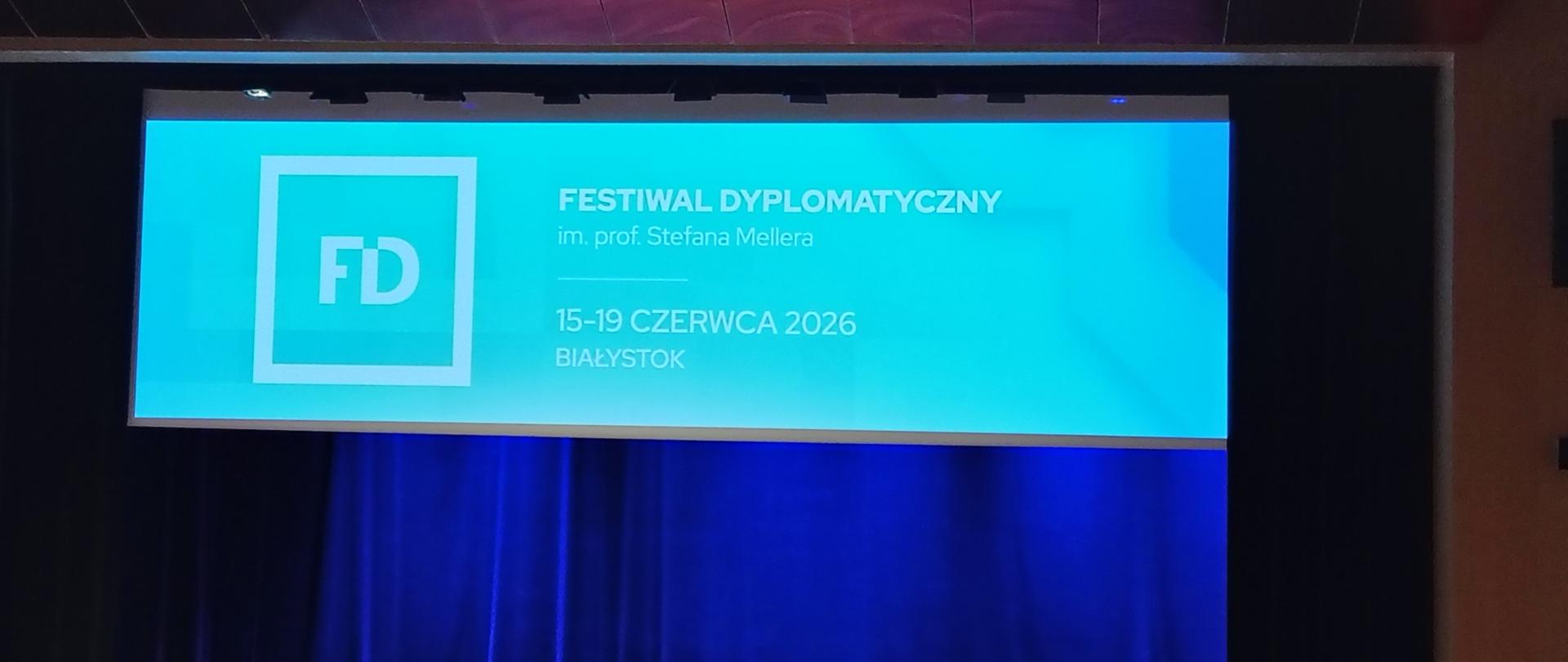 Scena z dużym ekranem, na którym wyświetlana jest prezentacja, otoczona przez publiczność. 