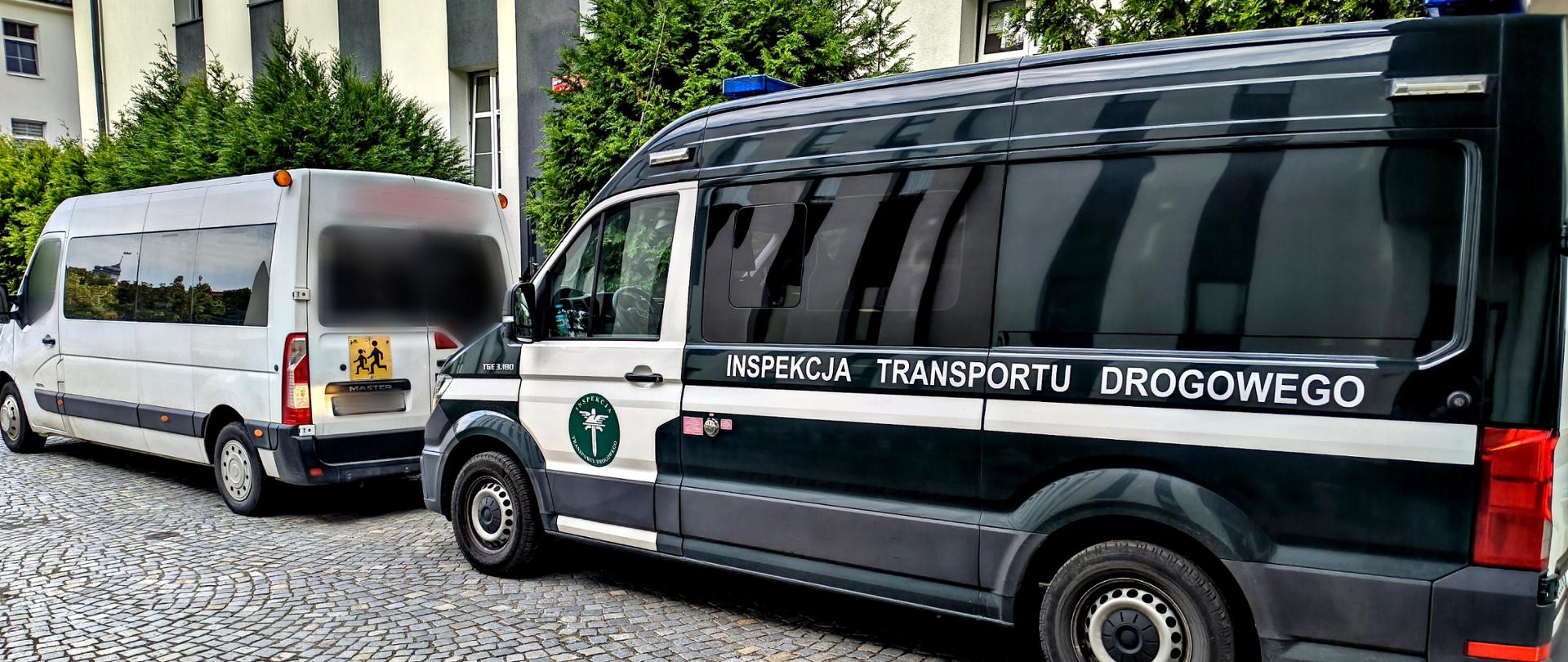 Dwa pojazdy stoją jeden za drugim na kostce brukowej przed budynkiem. Na pierwszym planie, widoczny z boku, znajduje się ciemnozielony pojazd Inspekcji Transportu Drogowego z szerokim białym pasem, napisem „INSPEKCJA TRANSPORTU DROGOWEGO”, logo instytucji oraz niebieskimi lampami sygnalizacyjnymi na dachu. Przed nim zaparkowany jest biały autobus, widoczny od tyłu i z lewego boku. Na tylnych drzwiach autobusu znajduje się żółta tablica informująca o przewozie dzieci. Szyby oraz tablice rejestracyjne pojazdów zostały zamazane.