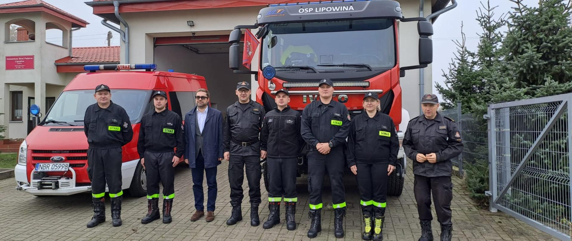Przy samochodach strażackich do zdjęcia pozują strażacy w czarnych mundurach oraz wójt. W tle remiza strażacka.