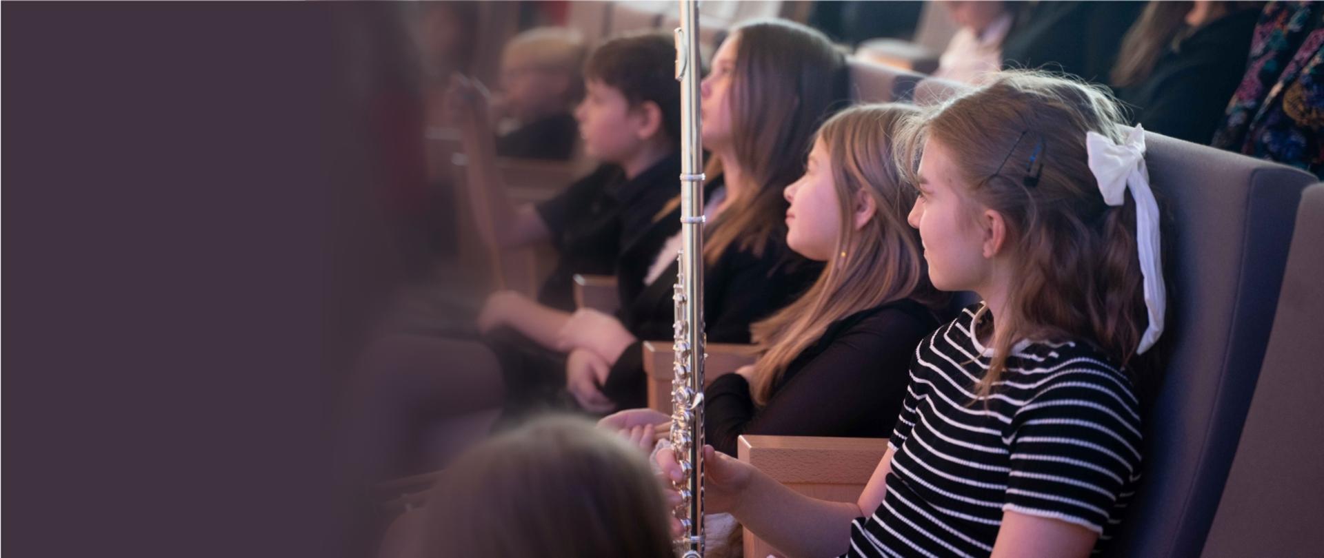 Zdjęcie przedstawia małą flecistkę zasiadającą na widowni w sali koncertowej ZPSM