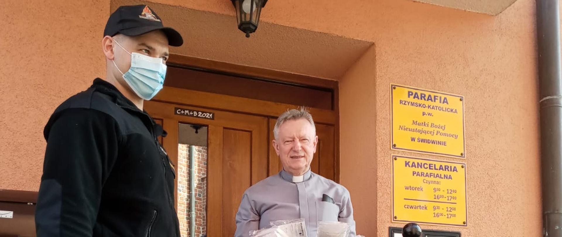 Zdjęcie przedstawia strażaka oraz księdza na tle parafii. strażak wręcza księdzu maseczki oraz płyn do dezynfekcji.
