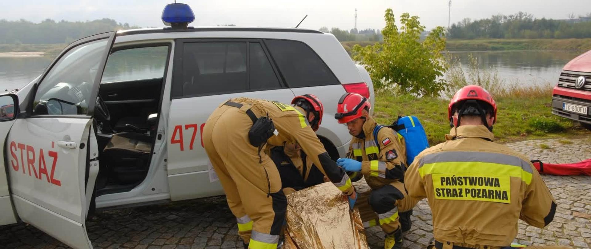 Strażacy Państwowej Straży Pożarnej udzielają kwalifikowanej pierwszej pomocy poszkodowanemu przy użyciu folii termicznej (NRC) oraz deski ortopedycznej w pobliżu akwenu wodnego.