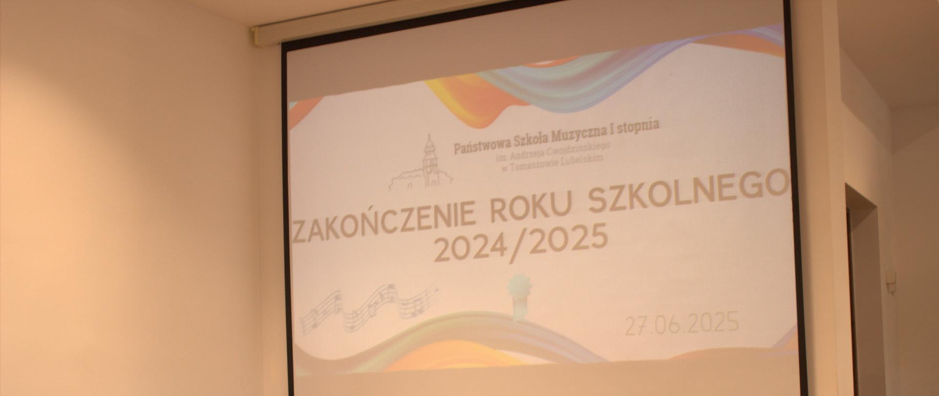 stół z nagrodami dla uczniów na zakończeniu roku szkolnego, powyżej projektor z wyświetlanym plakatem 