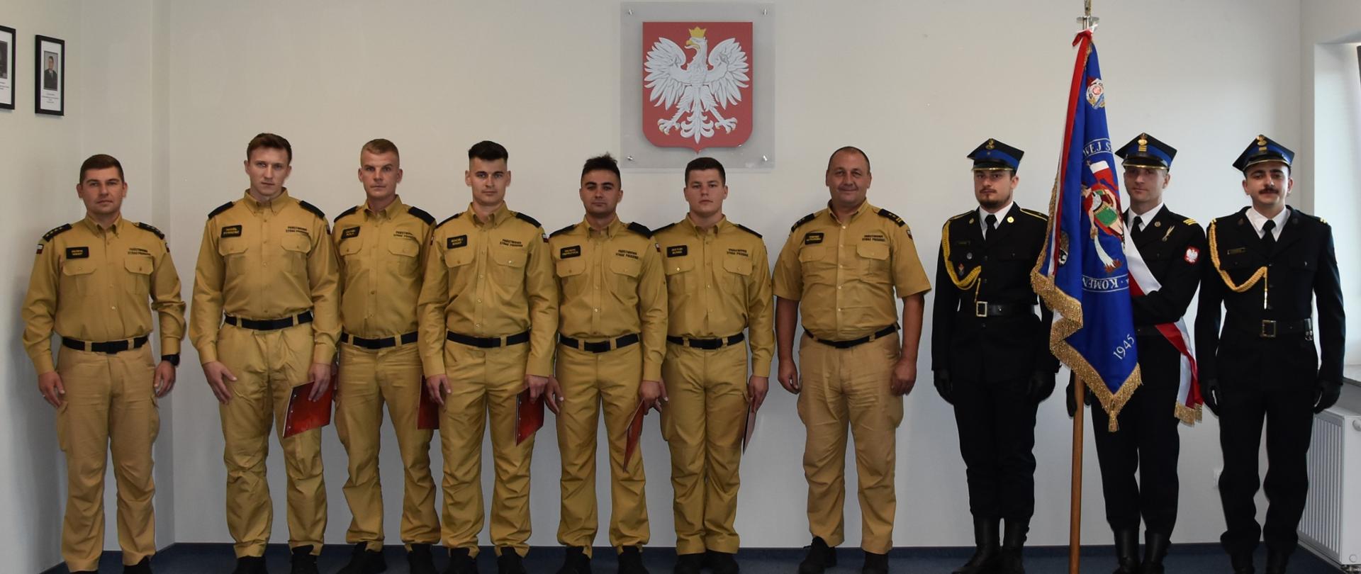 Na tle białej ściany stoją strażacy, którzy brali udział w uroczystym ślubowaniu. Siedmiu strażaków od lewej w mundurach piaskowych (spodnie i koszula). Trzech strażaków ze sztandarem w mundurach galowych. Na ścianie wisi godło.