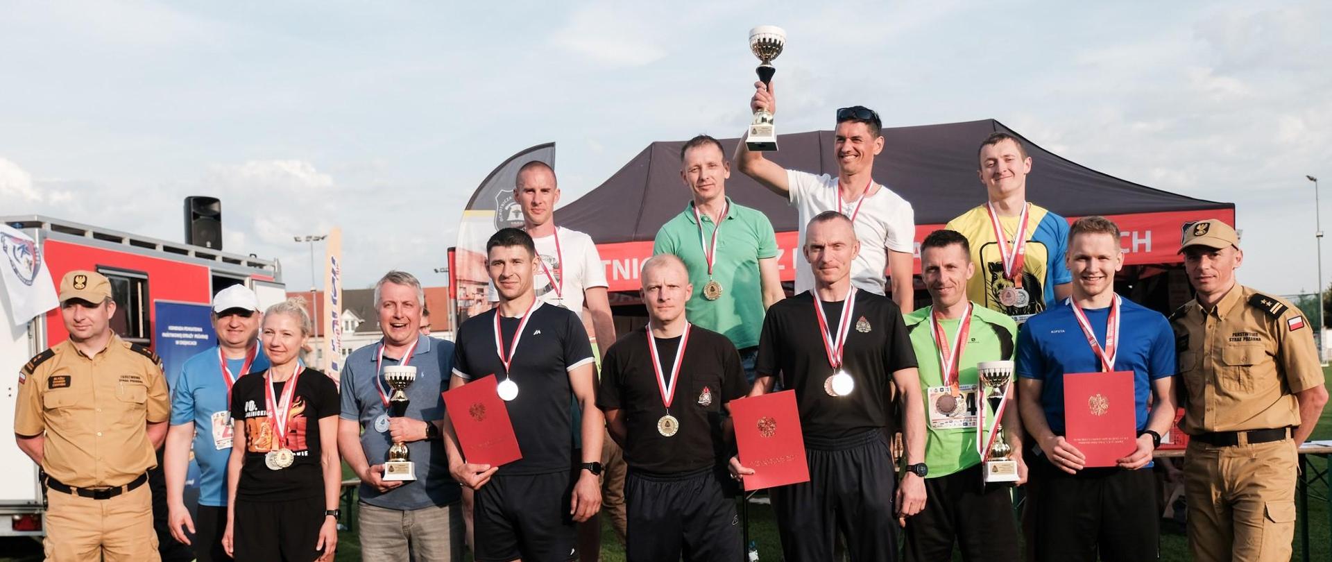 Strażacy stoją obok siebie wraz z medalami pucharami dyplomami po obu stronach stoją strażacy Państwowej Straży Pożarnej w mundurach koloru musztardowego za nimi ustawiony jest namiot ochotniczej straży pożarnej oraz samochód pożarniczy.