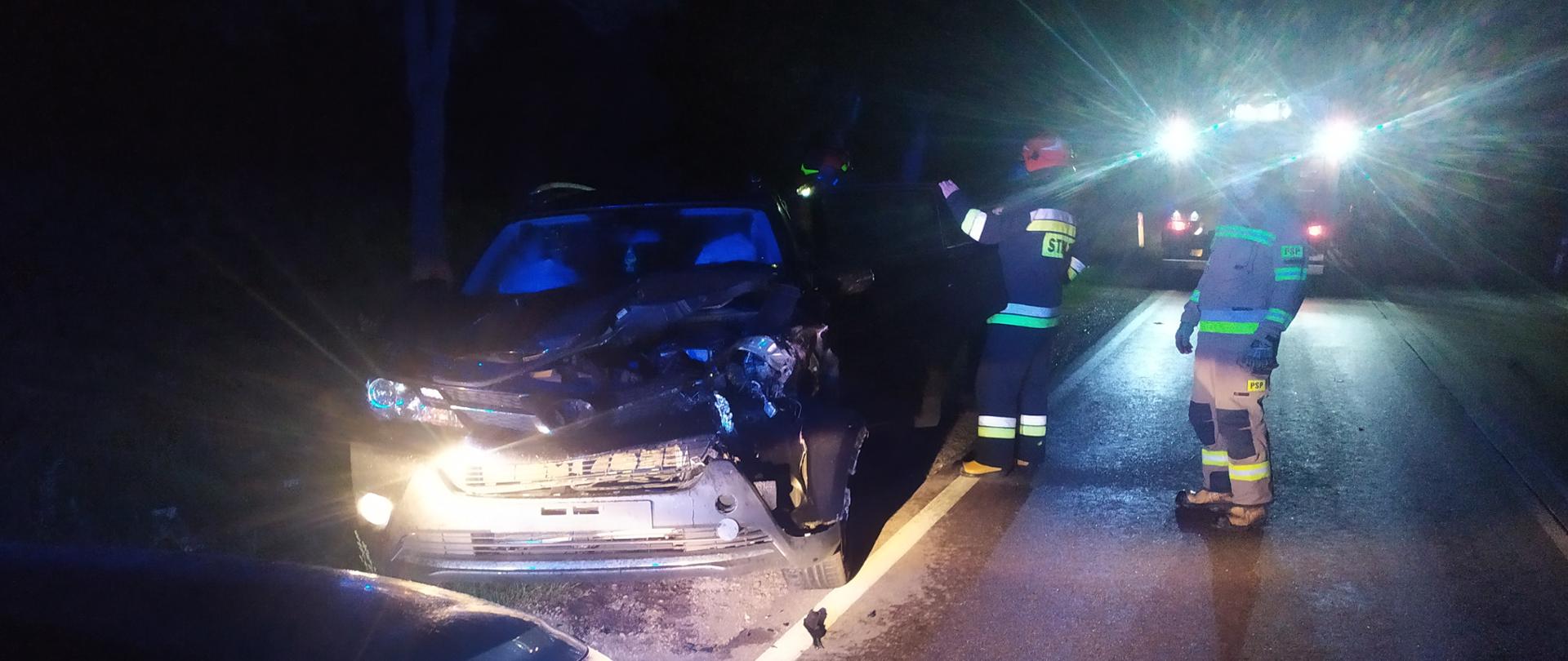 Samochód osobowy po zderzeniu z sarną. Uszkodzony przód samochodu. W tle strażacy i samochód strażacki.