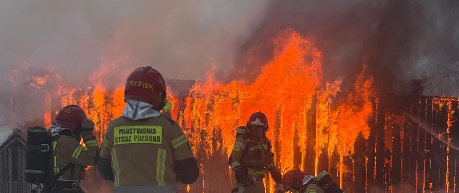 Zbliżenie na czterech strażaków pracujących przy rozwijaniu węży gaśniczych na śniegu przed płonącym budynkiem gospodarczym. Na plecach jednego ze strażaków widoczny jest odblaskowy napis „PAŃSTWOWA STRAŻ POŻARNA”, a na jego hełmie widnieje napis „RYSIEK”. W tle widać ścianę ognia trawiącą drewnianą konstrukcję obiektu.