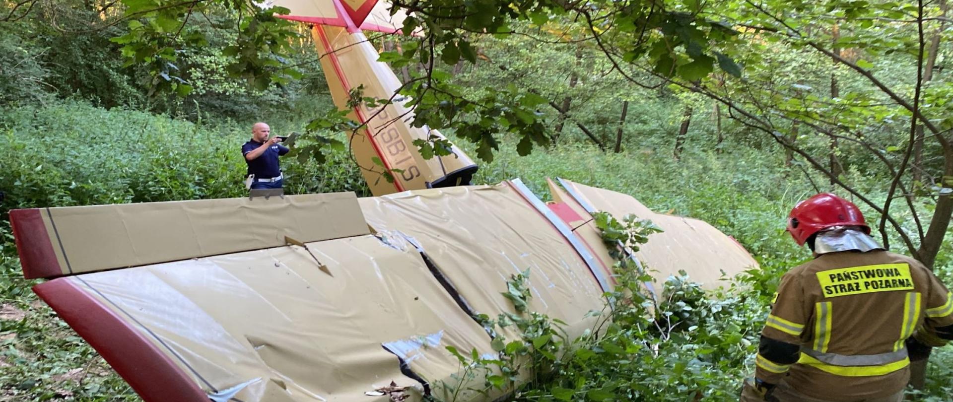 Zdjęcie przedstawia uszkodzony samolot ultralekki wbity dziobem w ziemie. Znajduje się w okolicy zakrzaczeń. Widać uszkodzenia skrzydeł samolotu. W tle pojawiają się funkcjonariusze służb działających na miejscu zdarzenia.