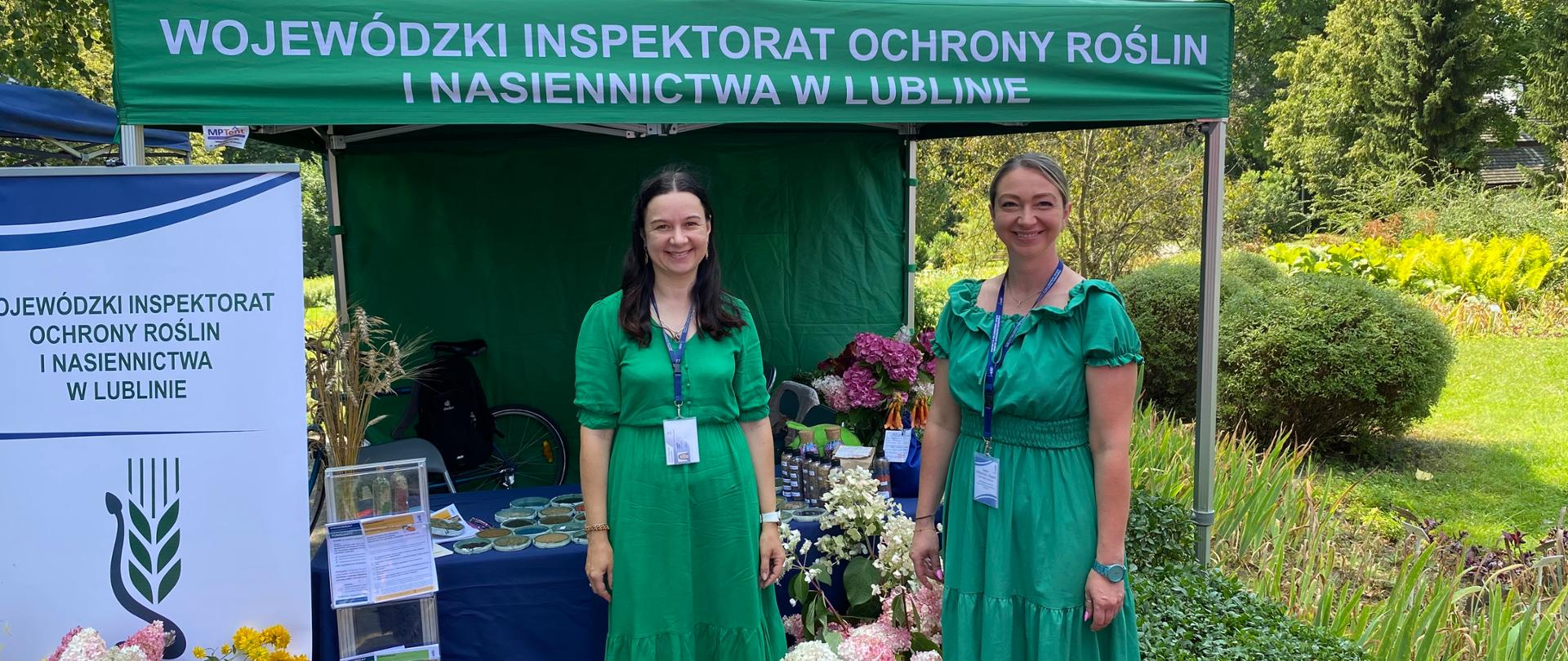 Dwie kobiety stojące przed namiotem. Obok stojak z ulotkami i stojak reklamowy. W tle stolik i kwiaty.