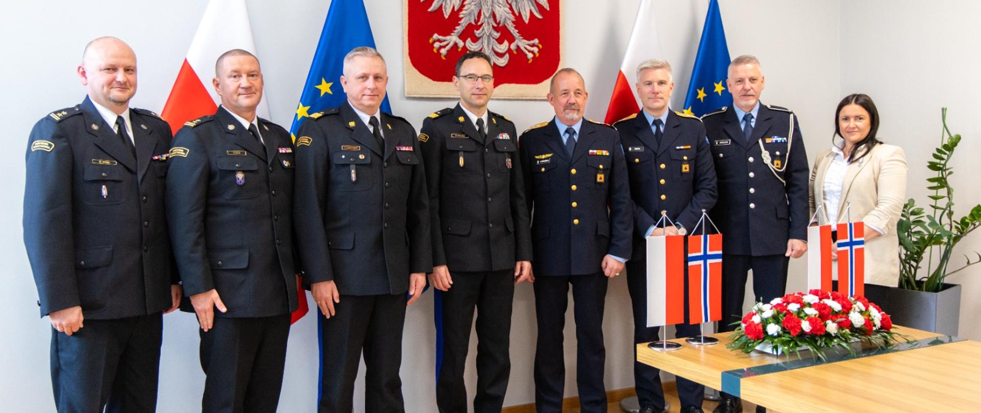 Grupa ludzi stojący w szeregu. W tle godło Polski.