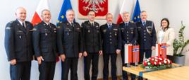 Grupa ludzi stojący w szeregu. W tle godło Polski.