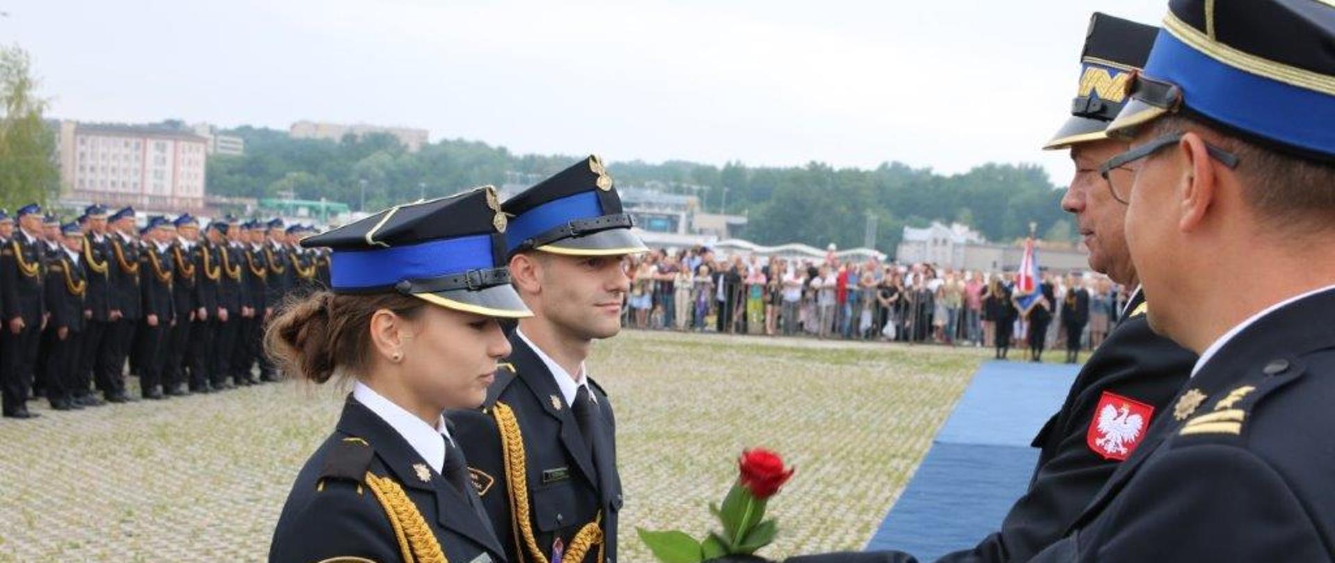 Zastępca KG PSP wręcza różę promowanej strażaczce za którą stoi strażak, w tle inni strażacy i rodziny