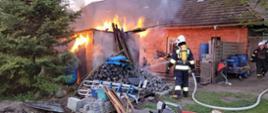 Widok na palący się budynek gospodarczy. Budynek mały, do około porozrzucane przedmioty. Jest dzień. Przy budynku strażacy podający prąd wody. W tle budynek mieszkalny z czerwonej cegły.
