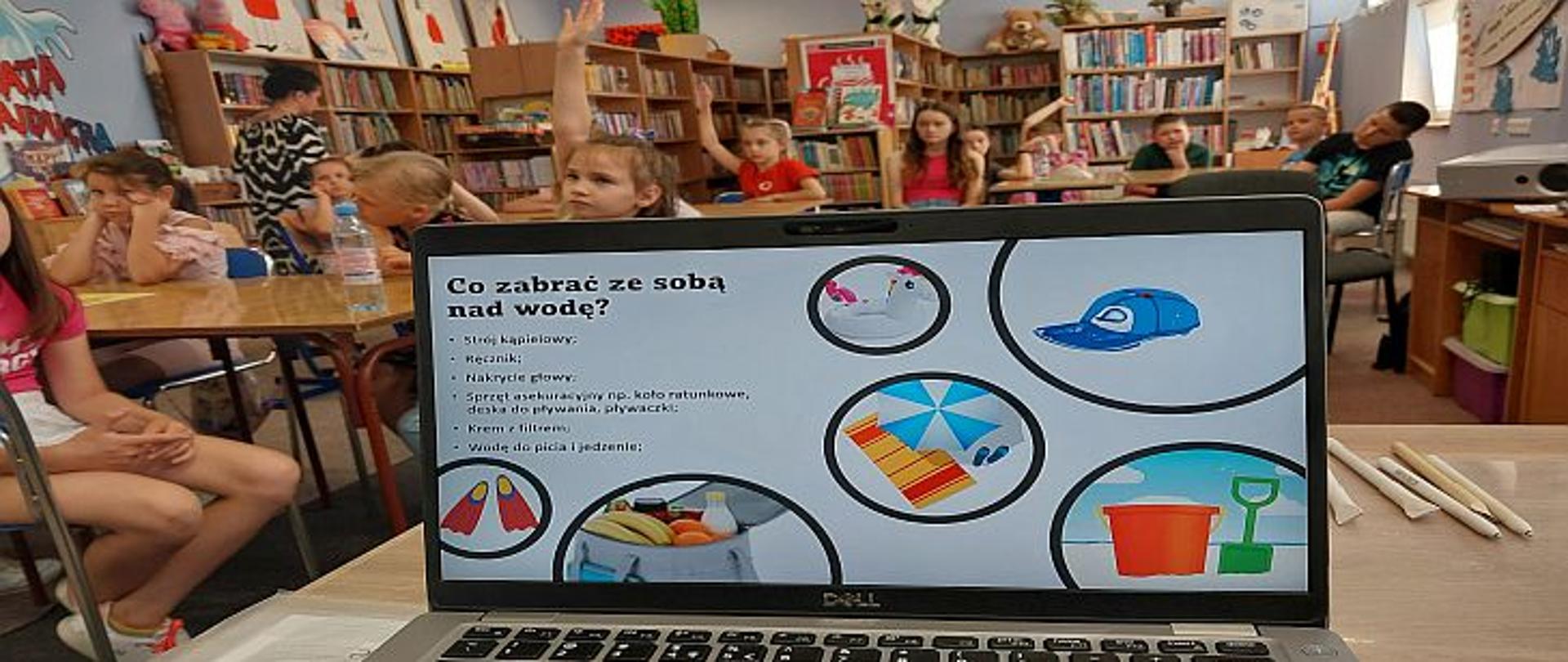 na pierwszym planie ekran monitora z napisem co zabrać ze sobą nad wodę, w tle twarze dzieci