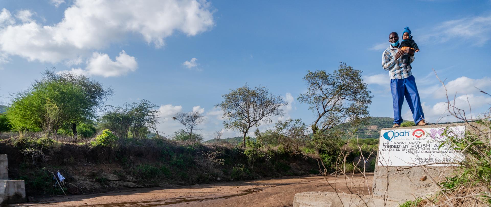 Mężczyzna z małym dzieckiem na rękach pozuje przy tamie piaskowej. W tle drzewa i odległe szczyty gór.