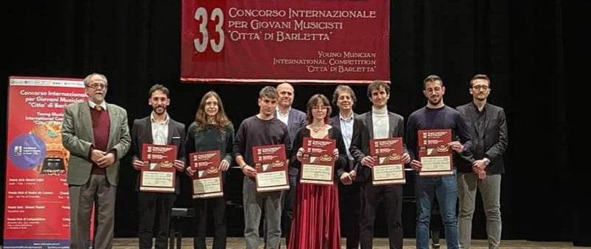 Zdjęcie kolorowe. Uczniowie z dyplomami oraz jurorzy na scenie. W tle baner konkursu (biały napis na czerwonym tle) o treści: "33 Concorso Internaziole per Giovani Musicisti "Citta di Barletta".