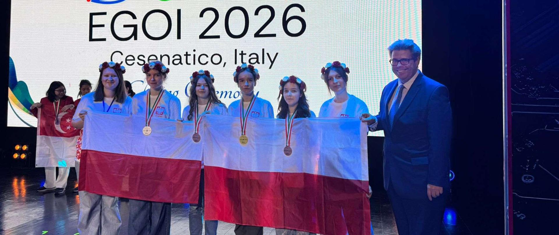 Grupa sześciu młodych kobiet w wiankach z kwiatów stoi na scenie, trzymając wspólnie dużą biało-czerwoną flagę Polski. Niektóre z nich mają na szyjach zawieszone medale. Po prawej stronie grupy stoi uśmiechnięty mężczyzna w okularach i ciemnoniebieskim garniturze. W tle znajduje się wielki, jasny ekran z logo wydarzenia oraz napisami „EGOI 2026” i „Cesenatico, Italy”. Po lewej stronie w głębi sceny widoczne są inne uczestniczki, z których jedna trzyma flagę Singapuru.