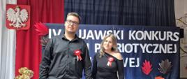 Zdjęcie przedstawia dwoje młodych ludzi, kobietę i mężczyznę, stojących obok siebie i pozujących do zdjęcia. Stoją oni na tle biało-czerwonych flag z godłem Polski oraz dużego, częściowo zasłoniętego transparentu. Scena sugeruje uroczystość patriotyczną lub szkolny konkurs o tej tematyce.