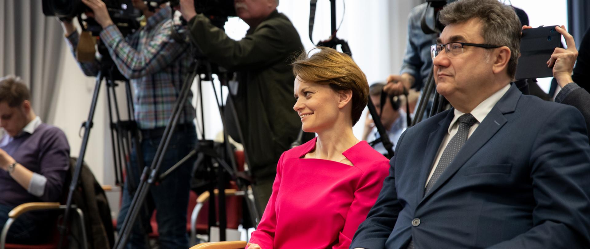 Minister Jadwiga Emilewicz i wiceminister Grzegorz Tobiszowski siedzą w pierwszym rzędzie podczas konferencji prasowej, w tle kamery. 
