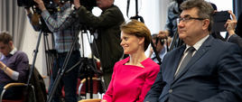Minister Jadwiga Emilewicz i wiceminister Grzegorz Tobiszowski siedzą w pierwszym rzędzie podczas konferencji prasowej, w tle kamery i dziennikarze.