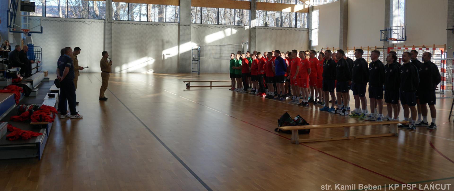 Zdjęcie przedstawia uczestników turnieju eliminacyjnego, którzy stoją na hali w dwuszeregu podzieleni na drużyny. Po lewej widoczny jest zastępca Komendanta Powiatowego PSP w Łańcucie , który przemawia podczas rozpoczęcia turnieju. 