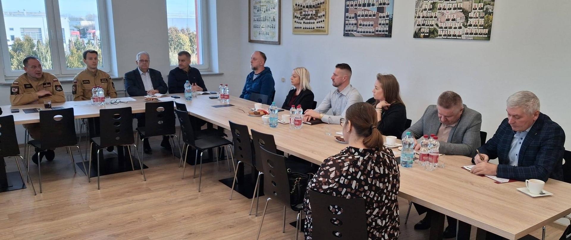 Na zdjęciu widać salę konferencyjną oraz uczestników spotkania. Osoby siedzą po obu stronach stołu, część z nich notuje, inni słuchają osoby zabierającej głos. Na ścianie w tle znajdują się plansze ze zdjęciami oraz symbole związane z Państwową Strażą Pożarną.