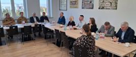 Na zdjęciu widać salę konferencyjną oraz uczestników spotkania. Osoby siedzą po obu stronach stołu, część z nich notuje, inni słuchają osoby zabierającej głos. Na ścianie w tle znajdują się plansze ze zdjęciami oraz symbole związane z Państwową Strażą Pożarną.