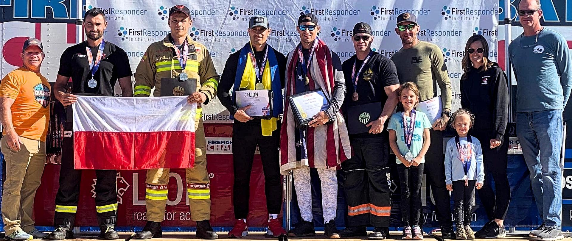 Jedenastoosobowa grupa w tym dwójka dzieci stojąca na scenie. Zdobywcy pierwszego miejsca trzymają w rekach pamiątkowe dyplomy, na szyjach wszystkich widoczne są zdobyte medale. Zdobywcy drugiego miejsca - dwóch mężczyzn trzyma w reku polską flagę. W tle uczestników baner reklamowy mistrzostw. 