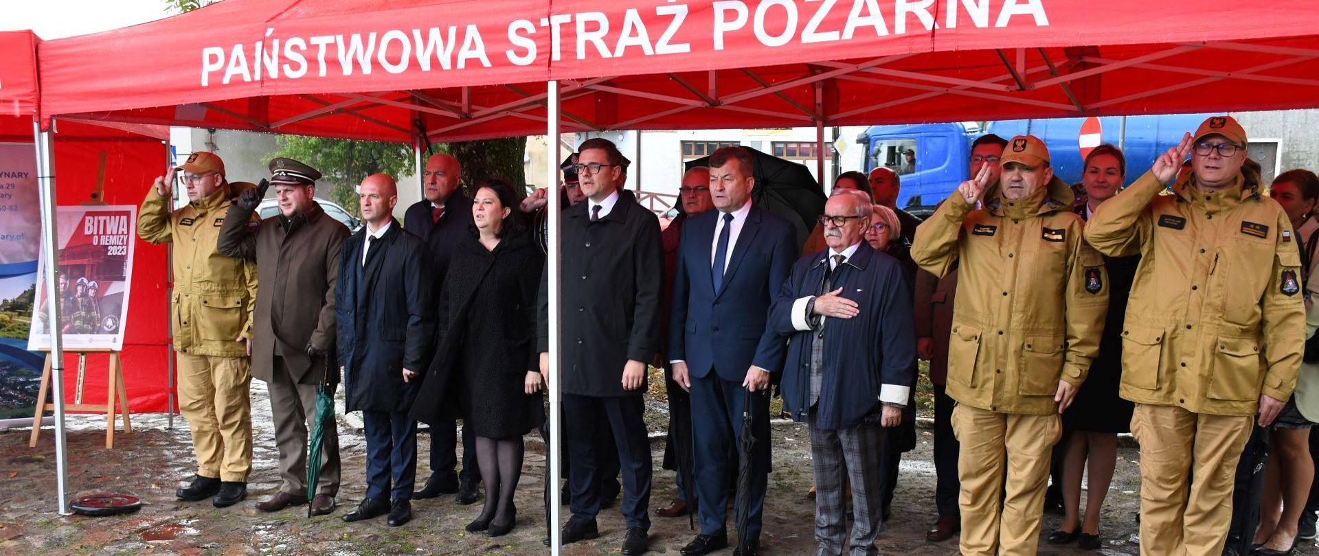 Pod czerwonym namiotem państwowej Straży Pożarnej stoją przedstawiciele rządu, samorządu oraz strażacy.