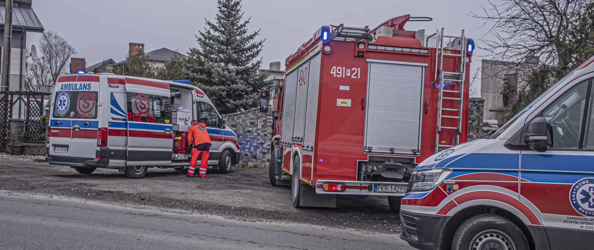Ratownik wybiera sprzęt z karetki. Na miejscu dwie karetki oraz wóz strażacki. Na zdjęciu widać ratownika który szykuje sprzęt medyczny. 