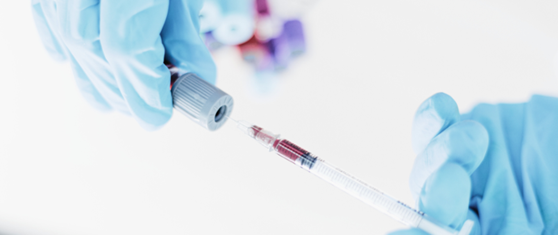 Zdjęcie przedstawia dwie dłonie w rękawiczkach które strzykawką pobierają próbkę z fiolki 