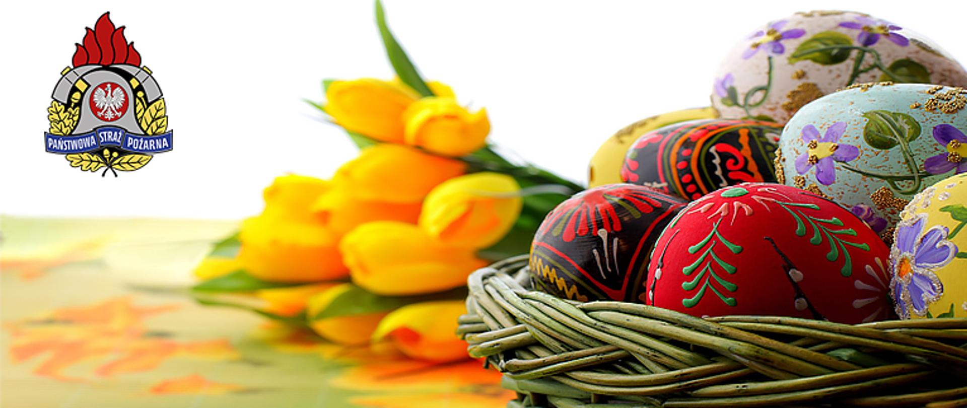 siedem pisanek znajduje się w wiklinowym koszyczku z prawej stronu zdjęcia w tle widoczne tulipany barwy żółtej, z lewej w górnym rogu strony logo Państwowej Straży Pożarnej a pod nim napis Komenda Powiatowa PSP w Łowiczu