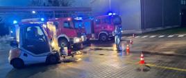 Na zdjęciu wózek widłowy w nim strażak ubrany w ubranie gazoszczelne za wózkiem stoją dwa samochody strażackie koloru czerwonego z włączonymi światłami przed nim stoi drugi strażak w ubraniu gazoszczelnym koloru niebieskiego przy nim widać rozstawione pachołki koloru czerwono białego w oddali szara hala oraz oświetlenie z lampy 