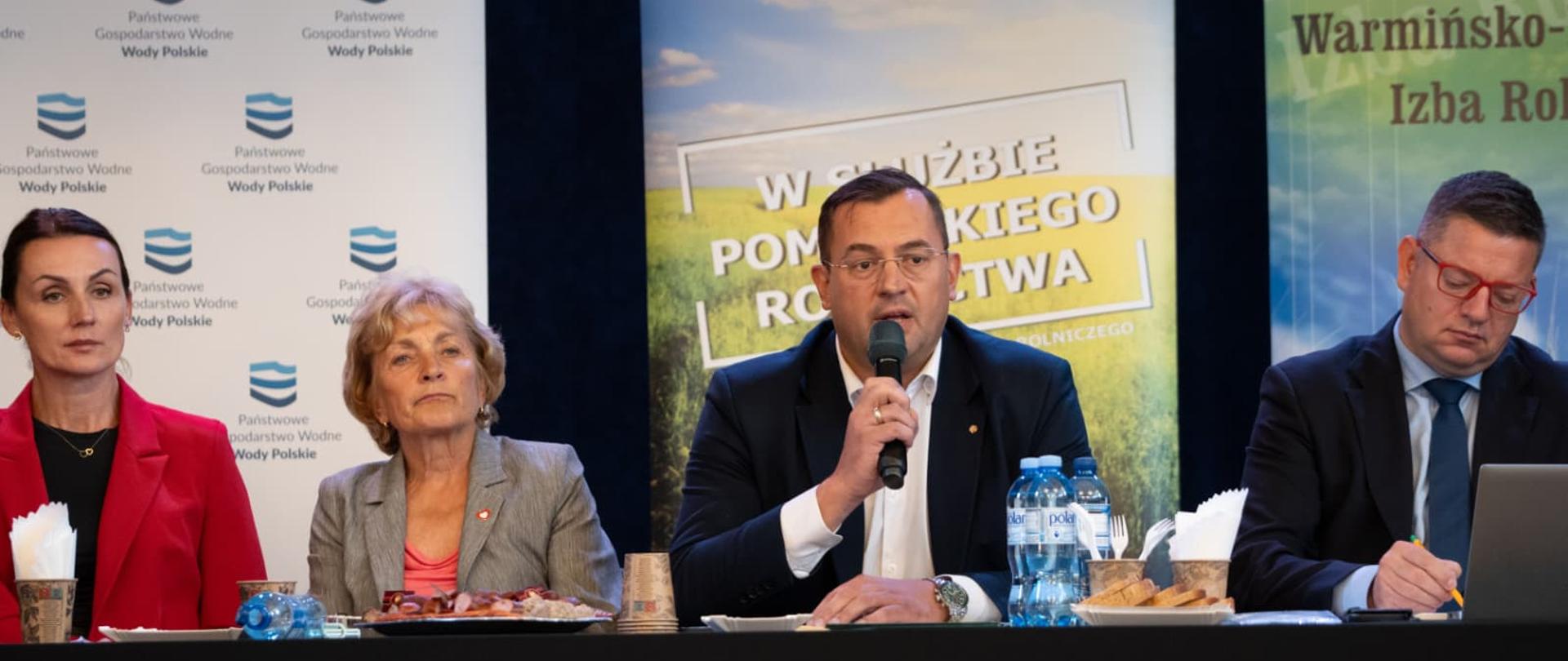 kilka osób siedzi za stołem prezydialnym, jeden z mężczyzn trzyma mikrofon