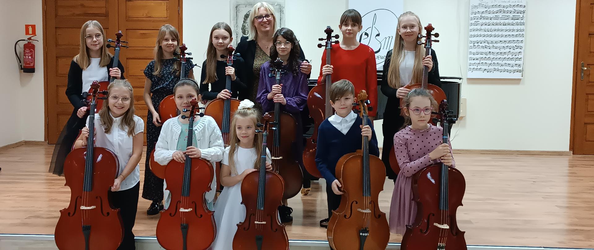Zdjęcie przedstawia uczniów klasy wiolonczeli Pani Urszuli Właźniak stojących z wiolonczelami na scenie i przed sceną. W tle po prawej stronie widać drewniane drzwi. Po lewej stronie widać grafikę czarno białą z partyturą. Za grupą wiolonczel widać grafikę z dawnym logo szkoły. Nad grafikami wisi Godło Polski.