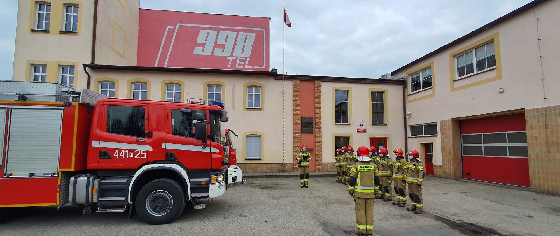 Na zdjęciu po prawej widoczni strażacy ubrani w piaskowe ubranie specjalne i czerwone hełmy podczas zbiórki. Po lewej stronie widoczna przednia część ciężarowego czerwono – białego samochodu gaśniczego. W tle budynek komendy z masztem, na którym powiewa flaga Polski.