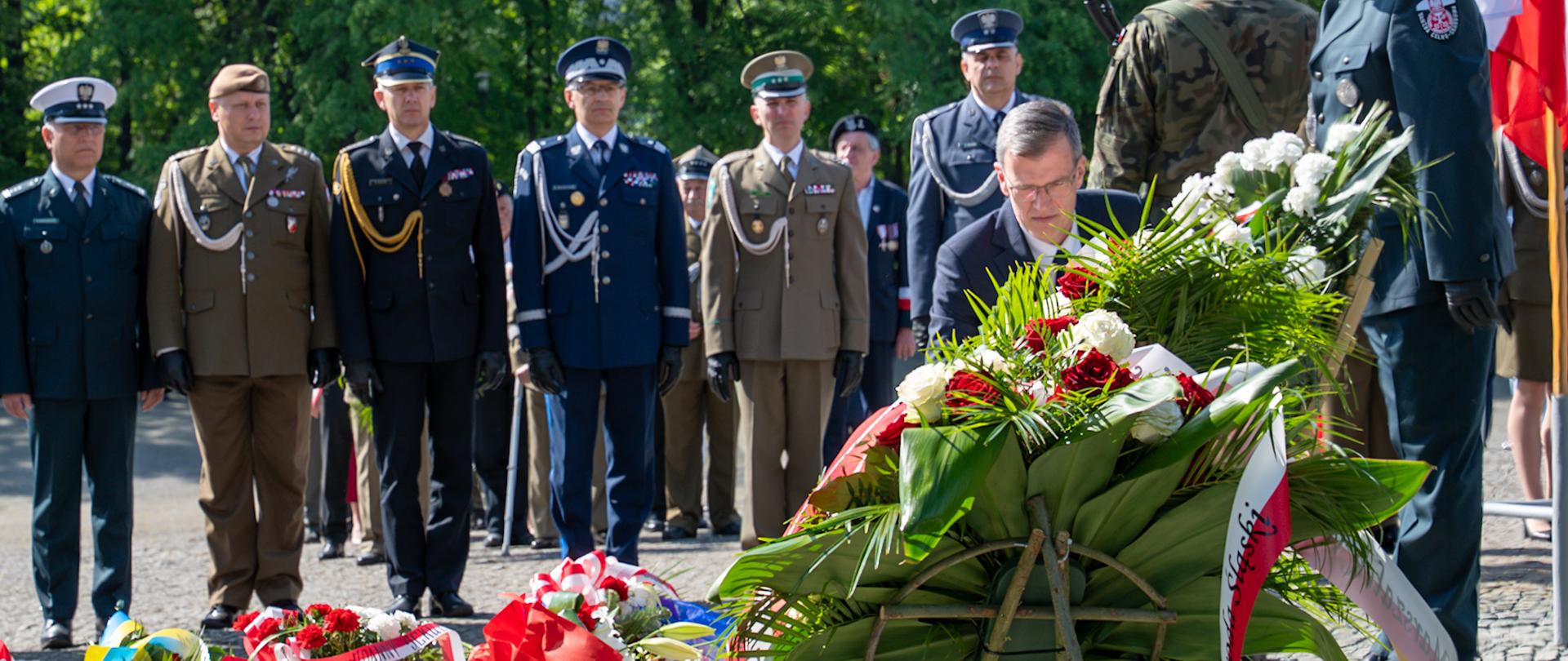 Na zdjęciu widoczna jest uroczystość upamiętniająca, podczas której składane są wieńce. Na pierwszym planie mężczyzna w garniturze, pochylony, układa duży, ozdobny wieniec z biało-czerwonych kwiatów (róż i goździków) oraz zielonych liści. Do wieńca przymocowana jest biało-czerwona wstęga z napisem „Bohaterom Śląskim”.
W tle stoi rząd oficerów Wojska Polskiego i innych służb mundurowych, w galowych mundurach różnych formacji – wojsk lądowych, lotnictwa, marynarki i policji. Wśród nich st. bryg. Roman Klecha zastepca śląskiego Komendanta Wojewódzkiego PSP. Za nimi widać drzewa w pełni zieleni, sugerujące wiosenną lub letnią porę. Obok wieńców leżą inne wiązanki kwiatów, wszystkie udekorowane biało-czerwonymi wstążkami, co wskazuje na charakter patriotyczny uroczystości.
