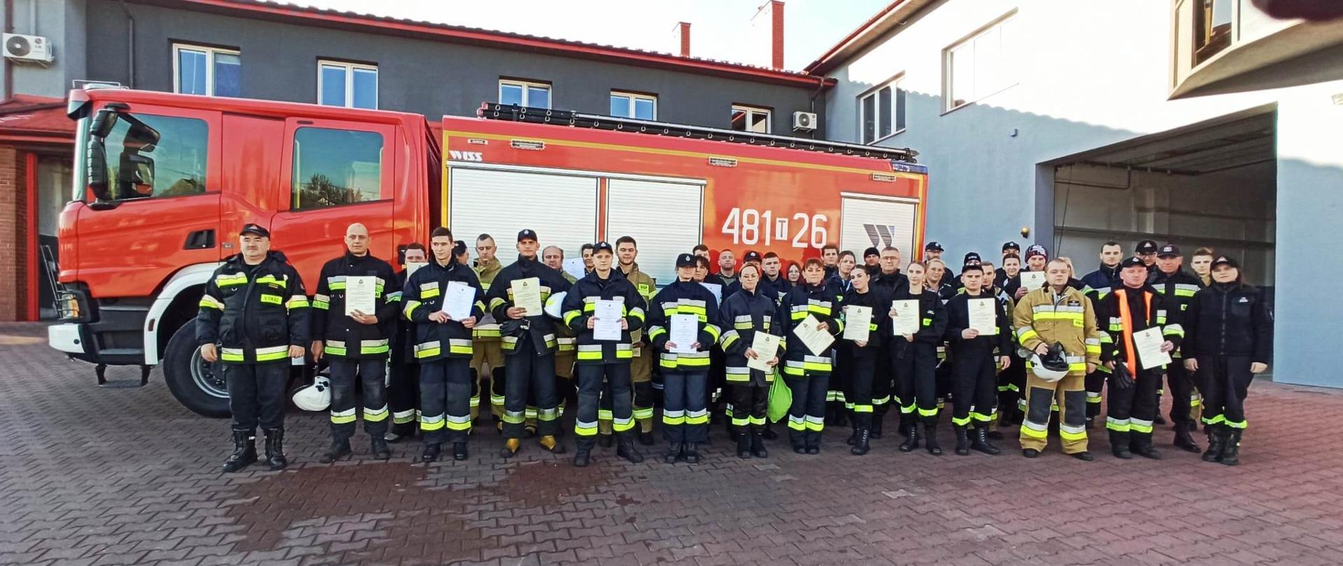 Druhowie OSP, którzy ukończyli kurs podstawowy pozują do wspólnego zdjęcia wraz z komisją egzaminacyjną. W tle widać samochód gaśniczy oraz budynek komendy powiatowej.