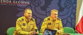 Na zdjęciu widać trzech strażaków podczas wideokonferencji, Trzy osoby siedzą przy stole do wideokonferencji. W tle flaga Polski po prawej stronie. W centrum i po lewej stronie ścianka koloru granatowego z orłem RP w odcieniu granatowym.