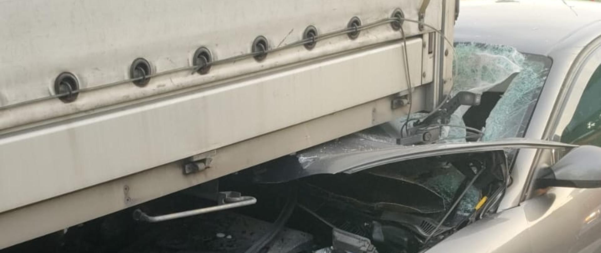 Na zdjęciu widoczny jest szary samochód osobowy marki Seat Ibiza, który wjechał w tył samochodu ciężarowego z zabudową plandekową. Przód samochodu osobowego jest poważnie uszkodzony – maska została zgnieciona i podniesiona, widoczne są uszkodzenia komory silnika, w tym akumulator. Szyba czołowa jest mocno pęknięta i rozbita po stronie kierowcy. Pojazdy znajdują się na pasie ruchu drogi asfaltowej, oznaczonej białymi liniami. W tle widać znak drogowy, ogrodzenie oraz fragment budynku. Zdarzenie miało miejsce w ciągu dnia, przy dobrej widoczności.