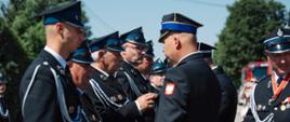 Zastępca Komendanta Powiatowego PSP przypina odznaczenie do munduru galowego strażaka OSP podczas oficjalnej uroczystości. W tle stoją pozostali strażacy w mundurach galowych