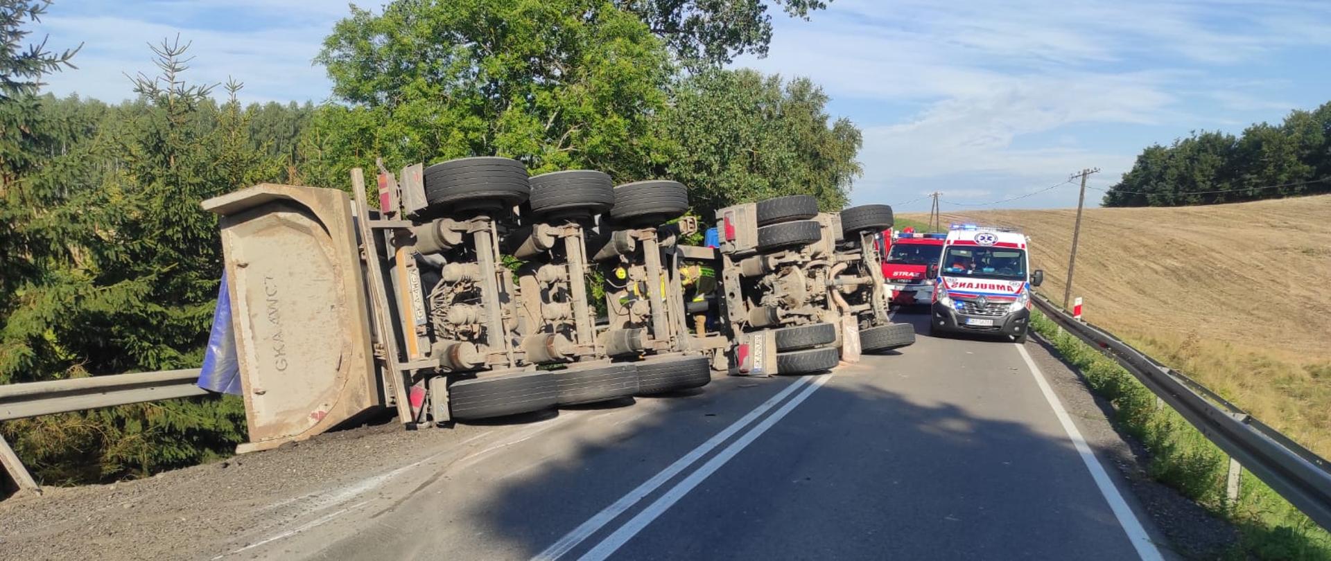 Na zdjęciu po lewej stronie widać przewróconą na bok przyczepę samochodu ciężarowego. W oddali po lewej stronie widać stojące samochody służb ratowniczych, zespołu ratownictwa medycznego oraz straży pożarnej.