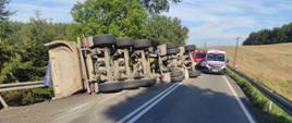 Na zdjęciu po lewej stronie widać przewróconą na bok przyczepę samochodu ciężarowego. W oddali po lewej stronie widać stojące samochody służb ratowniczych, zespołu ratownictwa medycznego oraz straży pożarnej.