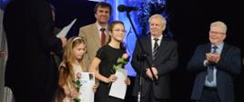 Na zdjęciu, na scenie widowiskowej JDK widnieje laureatka konkursu Julia Dąborowska, w
towarzystwie innej nagrodzonej uczennicy. Obok nagrodzonych uczennic widnieje jury
Ogólnopolskiego Konkursu Młodego Instrumentalisty im. Stefanii Wojtowicz w Jaśle.
Kolorystyka zdjęcia jest niebiesko-zielono-bezowo-czarna (autor zdjęcia: Katarzyna Pacwa).