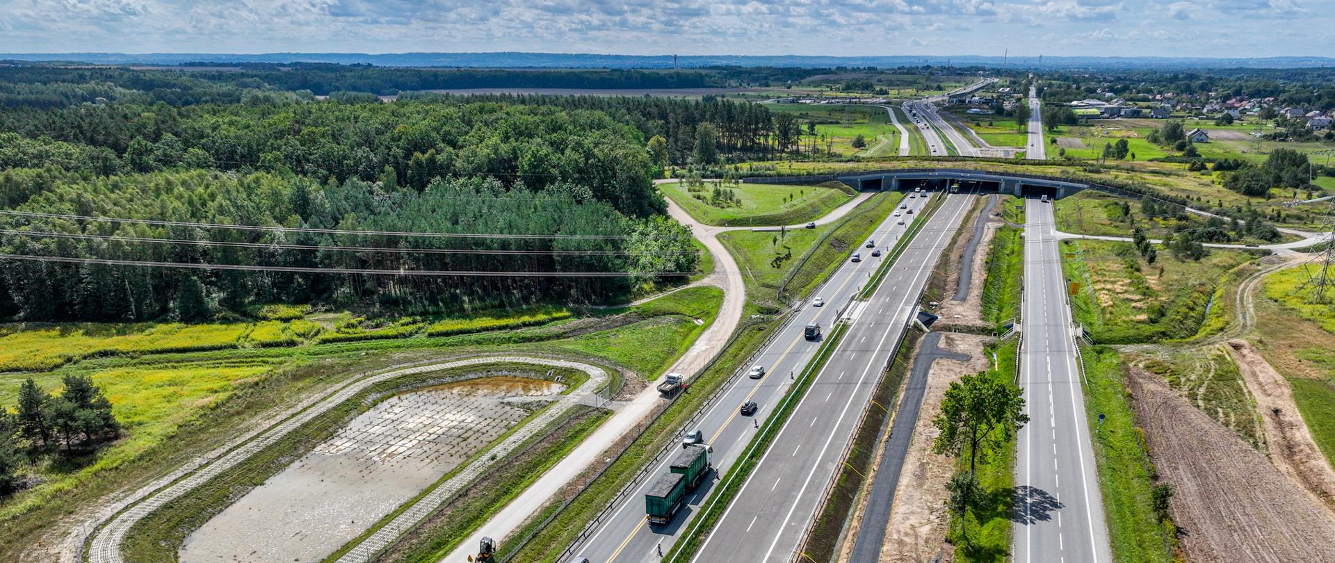 Widok z lotu ptaka na dwujezdniową drogę ekspresową biegnącą przez zielone tereny i las. Na jednej jezdni poruszają się samochody i ciężarówki, nad trasą znajduje się wiadukt, a obok widoczny jest zbiornik retencyjny, pola oraz rozproszona zabudowa.